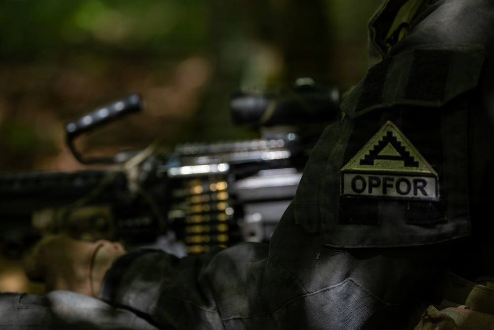 A close-up photograph of a military personnel wearing olive drab/woodland camouflage uniform with a visible 'OPFOR' (Opposing Force) patch and a unit insignia patch on the sleeve. In the background, a belt-fed machine gun with an ammunition belt is visible but out of focus. This appears to be a military training exercise scenario.