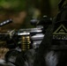 A close-up photograph of a military personnel wearing olive drab/woodland camouflage uniform with a visible 'OPFOR' (Opposing Force) patch and a unit insignia patch on the sleeve. In the background, a belt-fed machine gun with an ammunition belt is visible but out of focus. This appears to be a military training exercise scenario.