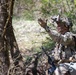 A heavily equipped soldier in multicam camouflage uniform is positioned in a wooded area, making a hand signal gesture. The soldier is wearing a helmet mounted with night vision goggles (flipped up), face paint, and carrying full combat load including body armor, rucksack, and an assault rifle. The setting appears to be a field training exercise or operational patrol in a temperate forest environment.