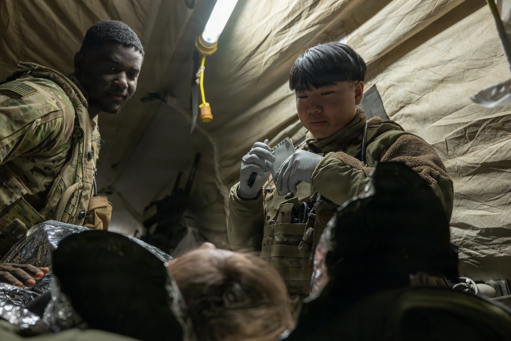 Two military personnel in MultiCam/OCP camouflage uniforms are conducting what appears to be a medical treatment or training exercise inside a field tent or shelter. One soldier wearing latex gloves is handling what appears to be a medical supply or dressing, while a second soldier observes. A patient or simulated casualty is visible in the foreground covered with what appears to be a thermal/emergency blanket.