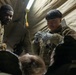 Two military personnel in MultiCam/OCP camouflage uniforms are conducting what appears to be a medical treatment or training exercise inside a field tent or shelter. One soldier wearing latex gloves is handling what appears to be a medical supply or dressing, while a second soldier observes. A patient or simulated casualty is visible in the foreground covered with what appears to be a thermal/emergency blanket.