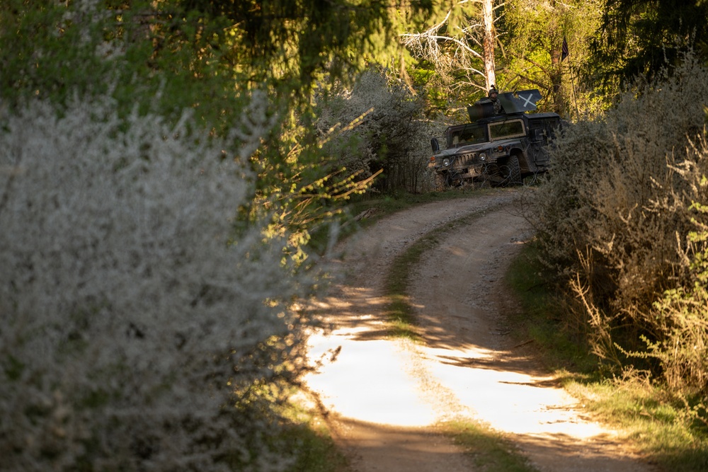 A military Humvee (HMMWV) is positioned off a narrow dirt road in a forested area, partially concealed among trees. The vehicle appears to have a marking or insignia visible on its side, and the scene suggests a tactical or training exercise in a woodland environment. The photograph is taken with a shallow depth of field, with blooming shrubs in the foreground rendered out of focus.