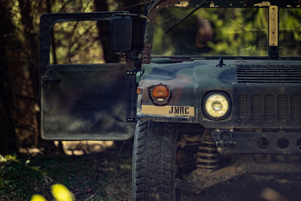 A military HMMWV (Humvee) is photographed in a forested environment, displaying camouflage paint and the stenciled marking 'JMRC' on its front bumper area. The vehicle's driver-side door is open, and a person is partially visible through the windshield. The image has a moody, dramatic quality with shallow depth of field and dappled forest lighting.