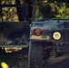 A military HMMWV (Humvee) is photographed in a forested environment, displaying camouflage paint and the stenciled marking 'JMRC' on its front bumper area. The vehicle's driver-side door is open, and a person is partially visible through the windshield. The image has a moody, dramatic quality with shallow depth of field and dappled forest lighting.