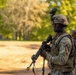 A U.S. Army soldier in full combat gear stands at alert in an outdoor training or operational environment. The soldier is wearing Operational Camouflage Pattern (OCP) uniform with a tactical helmet equipped with a night-vision device mount, and is holding an M4-style carbine rifle. Additional soldiers are visible in the background, suggesting a unit formation or patrol activity.