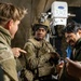 Three individuals are engaged in a discussion inside what appears to be a military field tent or command post. Two individuals are wearing military camouflage uniforms and tactical gear, while a third person in civilian-style clothing with glasses appears to be a civilian or interpreter. A medical or technical device labeled 'PhPRO' is visible in the background.