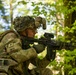 A U.S. Army soldier identified by the name tape 'SMITH' is positioned in a wooded area, aiming an assault rifle with an optic sight. The soldier is wearing full combat gear including OCP (Operational Camouflage Pattern) uniform, a helmet with camouflage netting and mounted night vision device brackets, and a tactical vest with equipment. The image appears to be taken during a field training exercise in a forest environment.