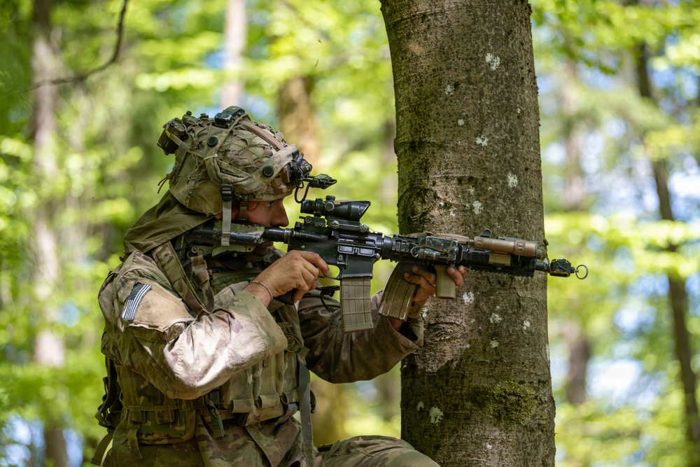 A soldier in full combat gear is positioned behind a tree in a forested environment, aiming a rifle in a tactical stance. The soldier is wearing MultiCam (OCP) pattern camouflage uniform with a US flag patch visible on the left shoulder. The setting appears to be a military training exercise or field operation in a deciduous woodland environment.