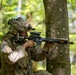 A soldier in full combat gear is positioned behind a tree in a forested environment, aiming a rifle in a tactical stance. The soldier is wearing MultiCam (OCP) pattern camouflage uniform with a US flag patch visible on the left shoulder. The setting appears to be a military training exercise or field operation in a deciduous woodland environment.