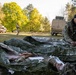Military personnel are conducting what appears to be a field medical training exercise in a wooded outdoor area. A soldier wearing a black beanie and multicam uniform attends to a simulated casualty wrapped in a mylar/emergency thermal blanket on the ground. Military vehicles including a HMMWV (Humvee) and a larger tactical truck are visible in the background.
