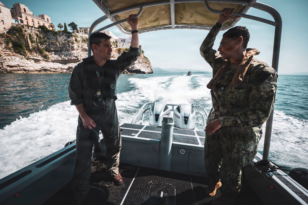 Two military personnel are standing on a moving rigid inflatable boat (RIB) on open water, both holding onto an overhead canopy frame for stability. One individual wears a dark flight suit with patches, while the other wears camouflage utilities with a tan vest. In the background, a rocky coastal cliff with buildings is visible, along with the boat's wake and a second vessel in the distance.