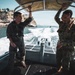 Two military personnel are standing on a moving rigid inflatable boat (RIB) on open water, both holding onto an overhead canopy frame for stability. One individual wears a dark flight suit with patches, while the other wears camouflage utilities with a tan vest. In the background, a rocky coastal cliff with buildings is visible, along with the boat's wake and a second vessel in the distance.
