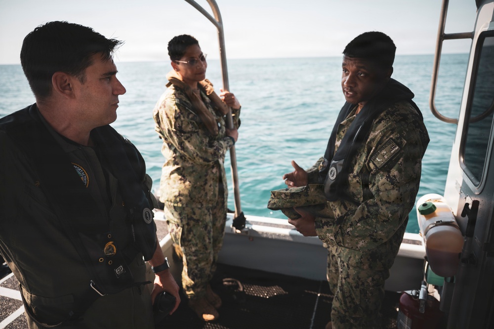 Three military personnel in camouflage uniforms are engaged in a conversation aboard a small patrol or utility boat at sea. Two of the individuals are wearing inflatable life vests over their uniforms, suggesting active maritime operations. The setting appears to be an open ocean environment with calm, blue-green water visible in the background.