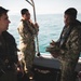 Three military personnel in camouflage uniforms are engaged in a conversation aboard a small patrol or utility boat at sea. Two of the individuals are wearing inflatable life vests over their uniforms, suggesting active maritime operations. The setting appears to be an open ocean environment with calm, blue-green water visible in the background.