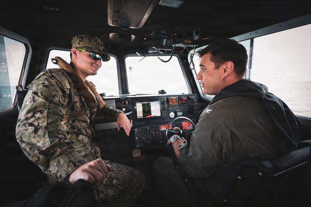 Two military personnel are seated inside the cockpit/helm area of a naval patrol vessel. One individual is wearing US Navy NWU Type III camouflage uniform with a cover (hat) and tactical vest, while the other is wearing an olive/sage flight suit. They appear to be engaged in conversation in a well-equipped helm station.