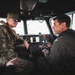 Two military personnel are seated inside the cockpit/helm area of a naval patrol vessel. One individual is wearing US Navy NWU Type III camouflage uniform with a cover (hat) and tactical vest, while the other is wearing an olive/sage flight suit. They appear to be engaged in conversation in a well-equipped helm station.