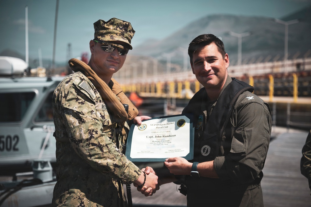 Two individuals are posing for a photo while jointly holding a certificate of achievement. The certificate is issued by the 'NAVND Guantanamo Harbor Patrol Unit' and is awarded to 'Capt. John Randazzo' as 'Honorary Coxswain.' The setting appears to be a naval or harbor facility, with a patrol boat numbered '502' visible in the background.