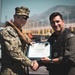 Two individuals are posing for a photo while jointly holding a certificate of achievement. The certificate is issued by the 'NAVND Guantanamo Harbor Patrol Unit' and is awarded to 'Capt. John Randazzo' as 'Honorary Coxswain.' The setting appears to be a naval or harbor facility, with a patrol boat numbered '502' visible in the background.