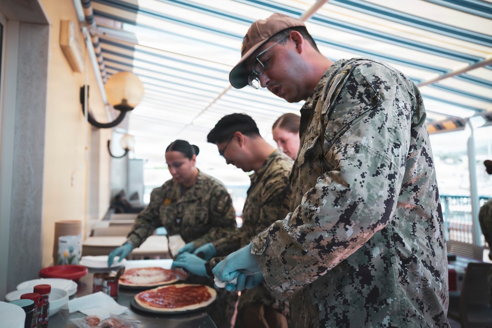 Several military personnel in digital camouflage uniforms are preparing pizzas at an outdoor or covered venue. They are wearing blue latex gloves and working with pizza dough and tomato sauce on a food preparation table. The setting appears to be a community or morale event where service members are engaged in food preparation.