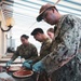 Several military personnel in digital camouflage uniforms are preparing pizzas at an outdoor or covered venue. They are wearing blue latex gloves and working with pizza dough and tomato sauce on a food preparation table. The setting appears to be a community or morale event where service members are engaged in food preparation.