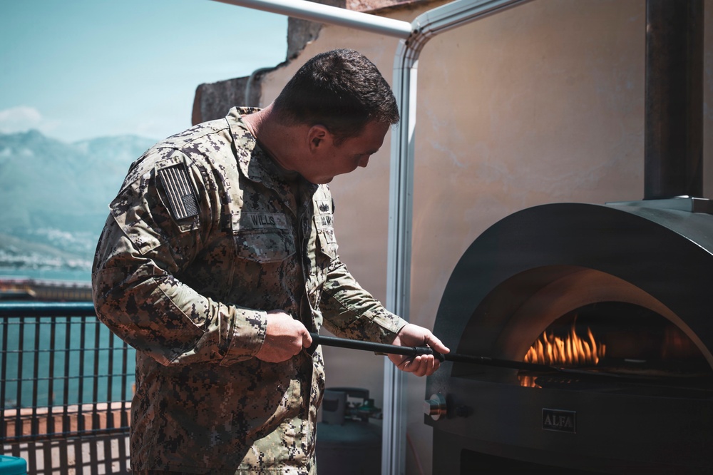 A U.S. Navy service member in camouflage uniform is using a long-handled peel or tool to tend to a wood-fired pizza oven on an outdoor terrace or rooftop. The individual's name tape reads 'WILLS' and an American flag patch is visible on the left sleeve. Mountains and a body of water are visible in the background, suggesting a Mediterranean or coastal location.