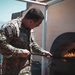 A U.S. Navy service member in camouflage uniform is using a long-handled peel or tool to tend to a wood-fired pizza oven on an outdoor terrace or rooftop. The individual's name tape reads 'WILLS' and an American flag patch is visible on the left sleeve. Mountains and a body of water are visible in the background, suggesting a Mediterranean or coastal location.