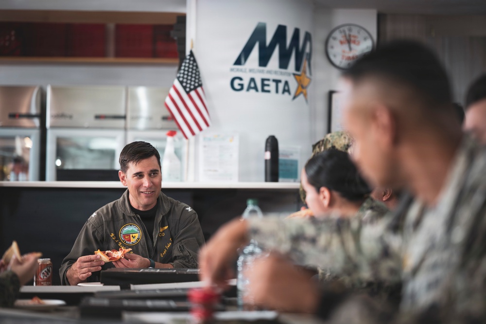 Military personnel are gathered at a dining facility located in Gaeta, Italy, as indicated by the MWR (Morale, Welfare, and Recreation) Gaeta banner on the wall. A senior officer in a flight suit with squadron patches is eating pizza and engaging in conversation with fellow service members. The setting appears to be a casual meal gathering at a U.S. Navy installation.
