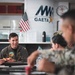 Military personnel are gathered at a dining facility located in Gaeta, Italy, as indicated by the MWR (Morale, Welfare, and Recreation) Gaeta banner on the wall. A senior officer in a flight suit with squadron patches is eating pizza and engaging in conversation with fellow service members. The setting appears to be a casual meal gathering at a U.S. Navy installation.