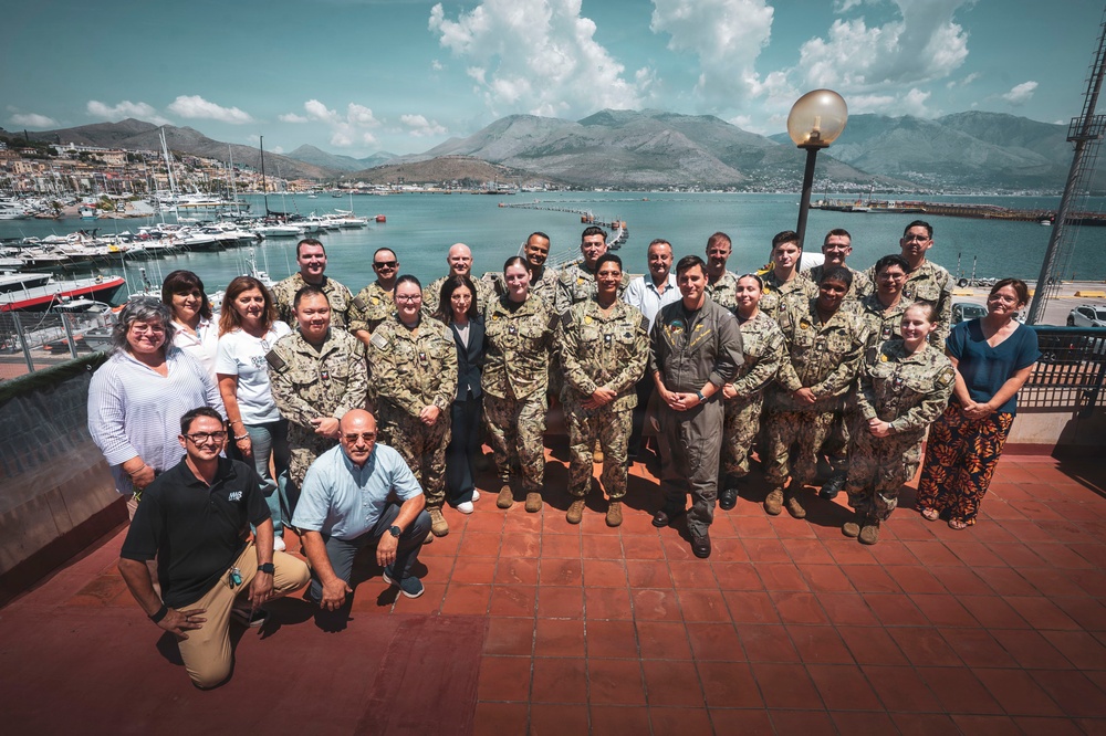 A large group of approximately 25-30 individuals poses for a formal group photograph on a waterfront terrace. The majority are wearing military camouflage uniforms, with several civilians in casual and business attire mixed among them. The backdrop features a Mediterranean-style harbor with moored boats, a coastal town, and mountain ranges under a partly cloudy sky.