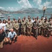 A large group of approximately 25-30 individuals poses for a formal group photograph on a waterfront terrace. The majority are wearing military camouflage uniforms, with several civilians in casual and business attire mixed among them. The backdrop features a Mediterranean-style harbor with moored boats, a coastal town, and mountain ranges under a partly cloudy sky.