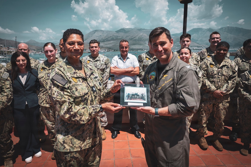 A formal military ceremony or exchange is taking place outdoors, likely at a naval installation near a coastal area with mountains in the background. Two senior military personnel — one in U.S. Navy digital camouflage (NWU) with the name tag 'Buchanan' and one in a flight suit — are jointly presenting or exchanging a framed photograph. A group of military personnel and at least one civilian stand behind them as witnesses.