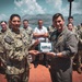 A formal military ceremony or exchange is taking place outdoors, likely at a naval installation near a coastal area with mountains in the background. Two senior military personnel — one in U.S. Navy digital camouflage (NWU) with the name tag 'Buchanan' and one in a flight suit — are jointly presenting or exchanging a framed photograph. A group of military personnel and at least one civilian stand behind them as witnesses.