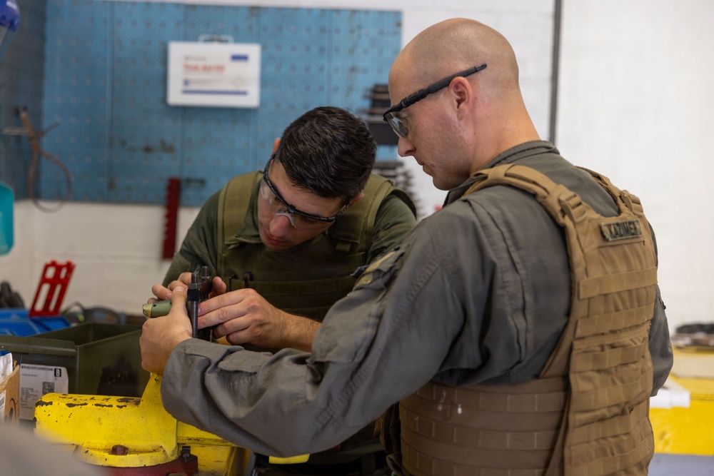 Two military personnel wearing tactical vests and safety glasses are working together on a piece of equipment in what appears to be a maintenance or ordnance workshop. Both individuals are focused on a task involving a small component or tool being handled at a workbench. The setting suggests a military maintenance facility with various tools and equipment visible in the background.