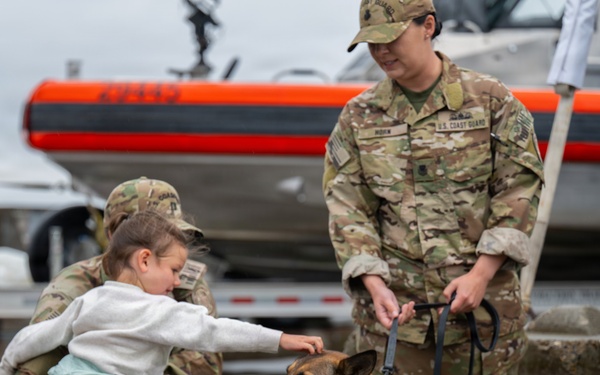Two U.S. Coast Guard personnel in OCP camouflage uniforms interact with a young child and a German Shepherd dog on a wet dock or pier. One service member is crouching and holding the child, who is reaching out to pet the dog, while the second service member stands holding the dog's leash. An orange and black rigid inflatable boat (RIB) is visible in the background.