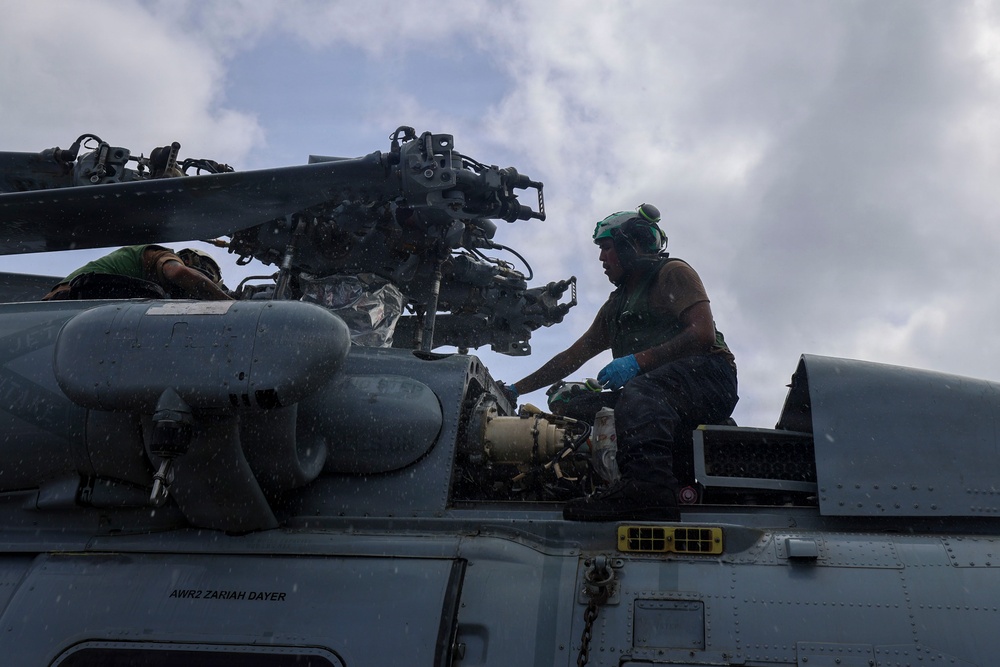 Two military personnel in green uniforms and helmets are performing maintenance work on top of a military helicopter. The work appears to be taking place outdoors under overcast, possibly rainy conditions. The text 'AWR2 ZARIAH DAYER' is visible on the fuselage of the aircraft.