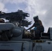 Two military personnel in green uniforms and helmets are performing maintenance work on top of a military helicopter. The work appears to be taking place outdoors under overcast, possibly rainy conditions. The text 'AWR2 ZARIAH DAYER' is visible on the fuselage of the aircraft.