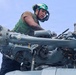 A military aviation mechanic or technician is performing maintenance work on what appears to be a helicopter rotor head assembly. The individual is wearing a green flight deck vest and a green helmet with hearing protection, indicating a naval aviation environment. The work involves close inspection or adjustment of the rotor hub mechanism.