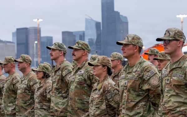 A formation of U.S. Coast Guard personnel standing at attention in OCP (Operational Camouflage Pattern) uniforms at what appears to be a waterfront ceremony or muster. The group includes both male and female service members, with a large urban skyline visible in the background across the water. The setting appears to be a pier or dock area under overcast, rainy conditions.