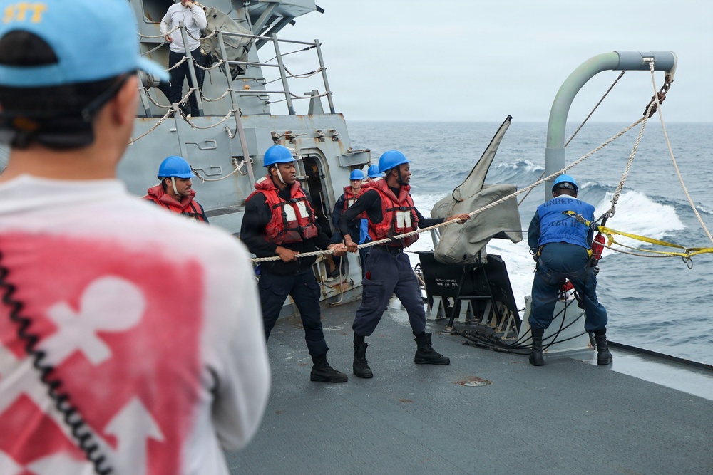 Naval personnel are conducting an underway replenishment (UNREP) operation on the deck of a warship at sea. Crew members wearing blue helmets and red life vests are hauling on a rope/line, while another crew member in blue coveralls marked 'UNREP' manages rigging near a replenishment rig. An observer in a red vest and blue cap watches from the foreground.