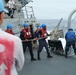 Naval personnel are conducting an underway replenishment (UNREP) operation on the deck of a warship at sea. Crew members wearing blue helmets and red life vests are hauling on a rope/line, while another crew member in blue coveralls marked 'UNREP' manages rigging near a replenishment rig. An observer in a red vest and blue cap watches from the foreground.