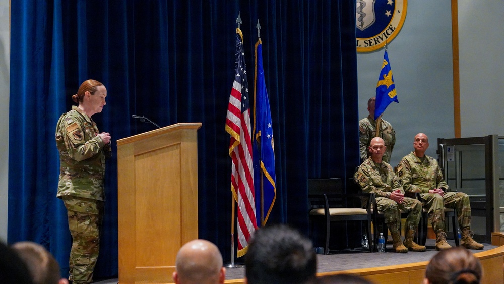 A military ceremony is taking place in a formal indoor venue, with a uniformed service member speaking at a wooden podium. Two seated military personnel observe from chairs on the stage, while a flag bearer stands behind them. The setting includes an American flag, a blue Air Force flag, and a partially visible official seal on the backdrop.
