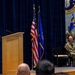 A military ceremony is taking place in a formal indoor venue, with a uniformed service member speaking at a wooden podium. Two seated military personnel observe from chairs on the stage, while a flag bearer stands behind them. The setting includes an American flag, a blue Air Force flag, and a partially visible official seal on the backdrop.