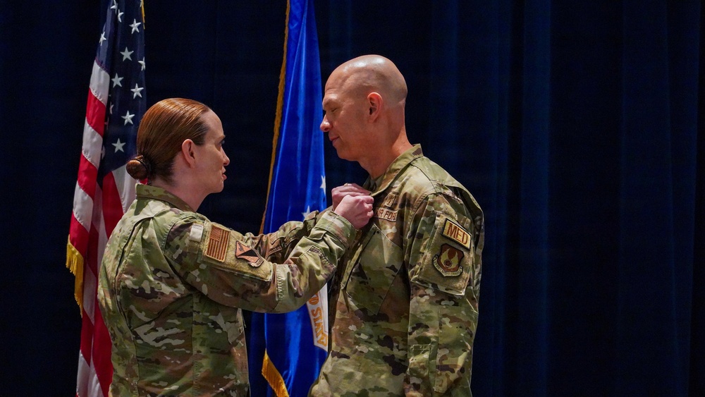 A military ceremony is taking place in which a female service member is pinning a medal or insignia onto a male service member's uniform. Both individuals are wearing OCP (Operational Camouflage Pattern) uniforms, and the male recipient has 'MED' and 'AIR FORCE' patches visible on his sleeve. The ceremony is conducted in front of an American flag and a U.S. Air Force flag.