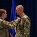 A military ceremony is taking place in which a female service member is pinning a medal or insignia onto a male service member's uniform. Both individuals are wearing OCP (Operational Camouflage Pattern) uniforms, and the male recipient has 'MED' and 'AIR FORCE' patches visible on his sleeve. The ceremony is conducted in front of an American flag and a U.S. Air Force flag.