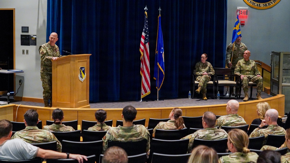 A military ceremony is taking place in an auditorium, likely a change of command or award ceremony for a U.S. Air Force unit. A uniformed officer is speaking at a podium on a raised stage while an audience of military personnel in OCP (Operational Camouflage Pattern) uniforms listens from seated rows. Two officers are seated on stage and one is standing holding a unit guidon/flag.