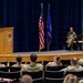 A military ceremony is taking place in an auditorium, likely a change of command or award ceremony for a U.S. Air Force unit. A uniformed officer is speaking at a podium on a raised stage while an audience of military personnel in OCP (Operational Camouflage Pattern) uniforms listens from seated rows. Two officers are seated on stage and one is standing holding a unit guidon/flag.