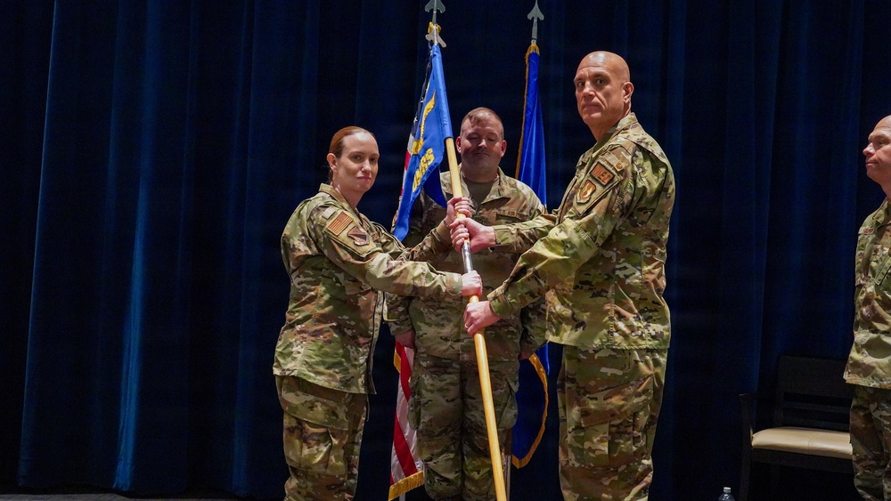 A military change of command ceremony is taking place on a stage with a dark blue curtain backdrop. Two service members in OCP (Operational Camouflage Pattern) uniforms are exchanging a unit guidon (flag on a staff), which is the traditional centerpiece of a change of command ceremony. Additional uniformed personnel are visible in the background and to the right.