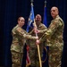 A military change of command ceremony is taking place on a stage with a dark blue curtain backdrop. Two service members in OCP (Operational Camouflage Pattern) uniforms are exchanging a unit guidon (flag on a staff), which is the traditional centerpiece of a change of command ceremony. Additional uniformed personnel are visible in the background and to the right.