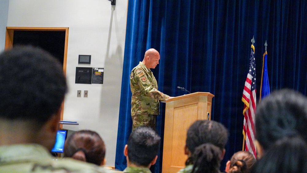 A bald military officer in OCP (Operational Camouflage Pattern) uniform is speaking at a wooden podium on a stage with a blue curtain backdrop. An American flag and what appears to be a service branch flag are displayed to the right of the podium. An audience of military and civilian personnel is seated in the foreground.