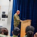 A bald military officer in OCP (Operational Camouflage Pattern) uniform is speaking at a wooden podium on a stage with a blue curtain backdrop. An American flag and what appears to be a service branch flag are displayed to the right of the podium. An audience of military and civilian personnel is seated in the foreground.